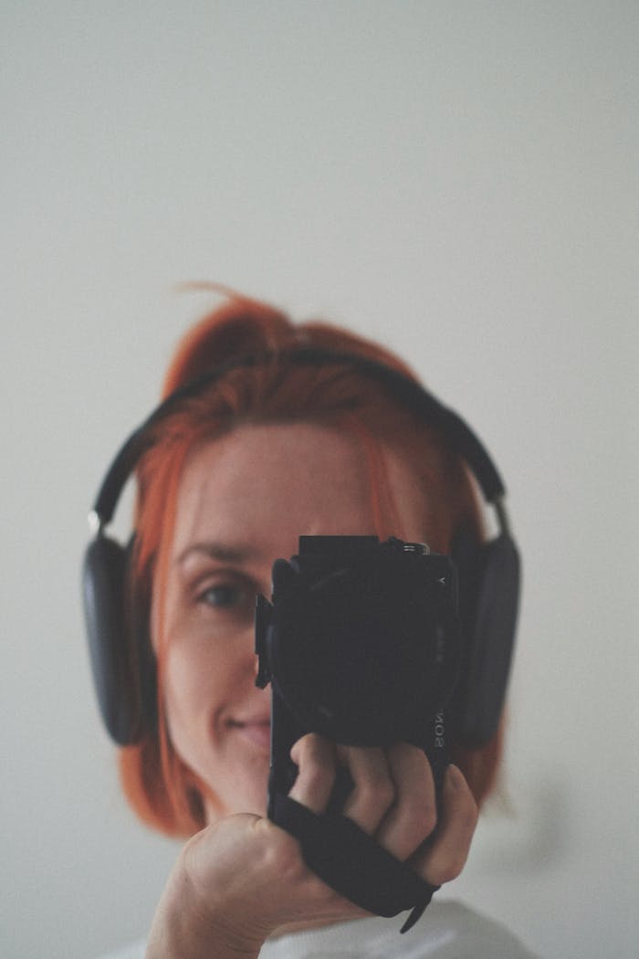 Redhead woman with headphones taking a self-portrait with a camera. Indoors, Gdansk, Poland.