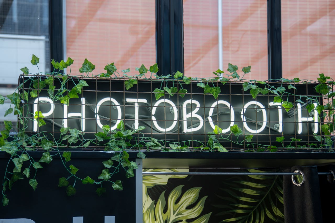 A photobooth sign with neon lights and greenery in an urban environment.